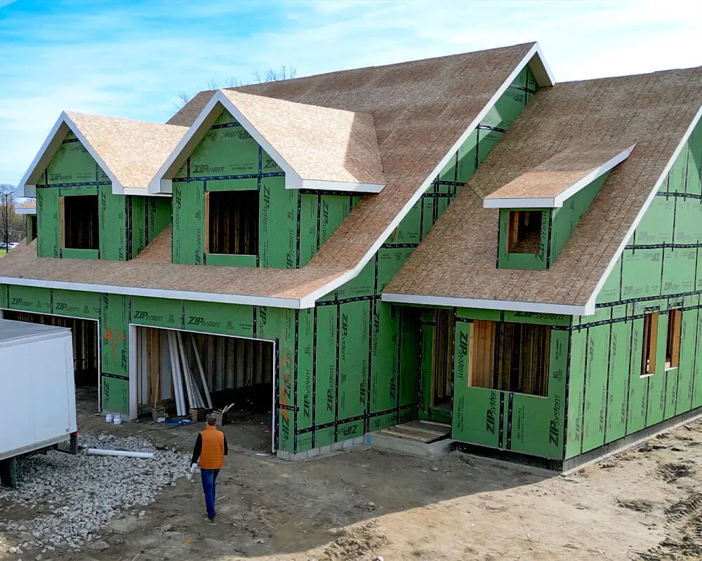 Exterior view of a two-story home under construction wrapped in green ZIP System sheathing, with a worker walking toward the entrance and a blue sky overhead.