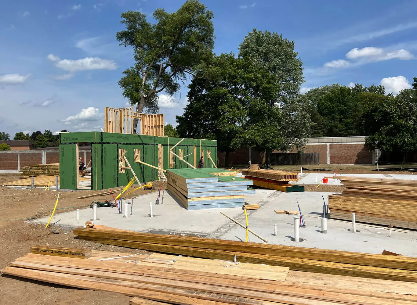 An active residential construction site showing partially framed walls with ZIP System sheathing, stacked lumber and building materials on a concrete foundation, surrounded by mature trees under a sunny sky.