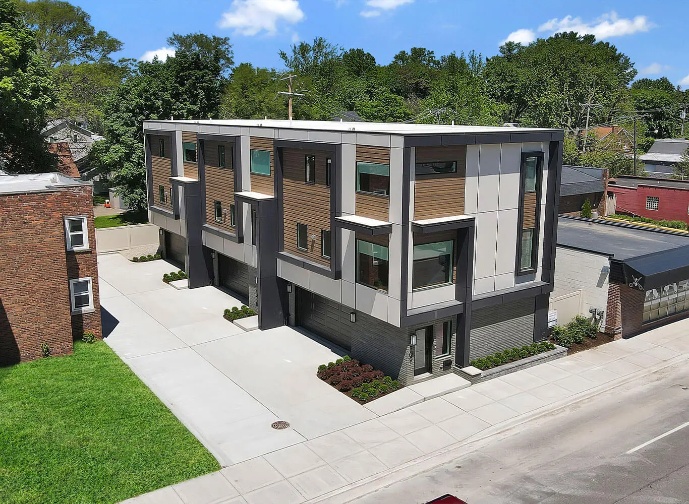 Aerial view of a modern three-story multi-unit building with wood and panel facade, dark trim, individual garage bays, and professional landscaping on a corner lot.