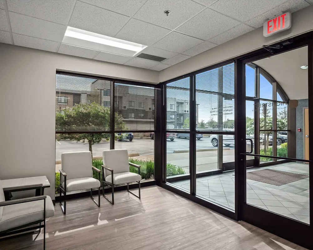 Interior lobby of a modern commercial office building with floor-to-ceiling windows, light wood flooring, contemporary seating, and glass entry doors.