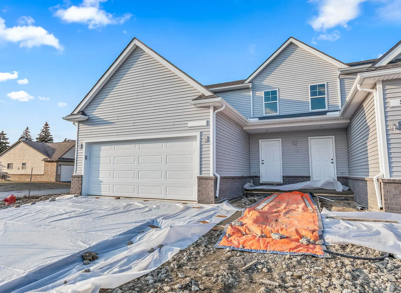Newly constructed townhome under construction with gray vinyl siding, white garage door, and exposed gravel and construction tarps in the foreground on a clear winter day.