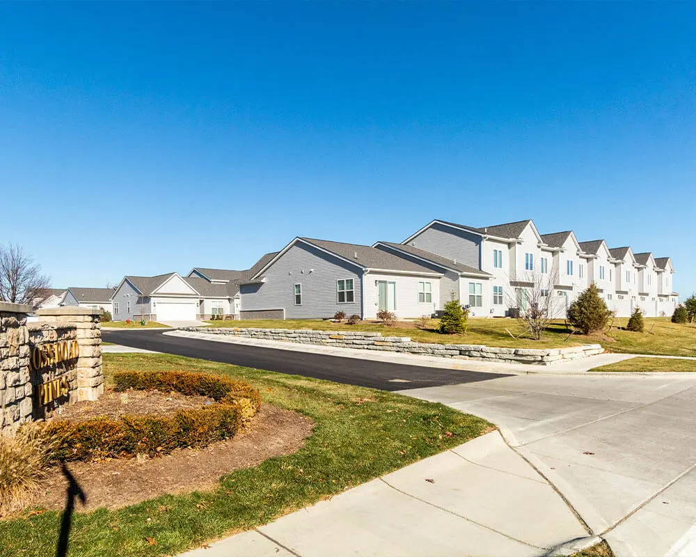 Entrance to a completed residential subdivision featuring a stone monument sign, rows of gray two-story townhomes, and well-maintained lawns along a freshly paved road.