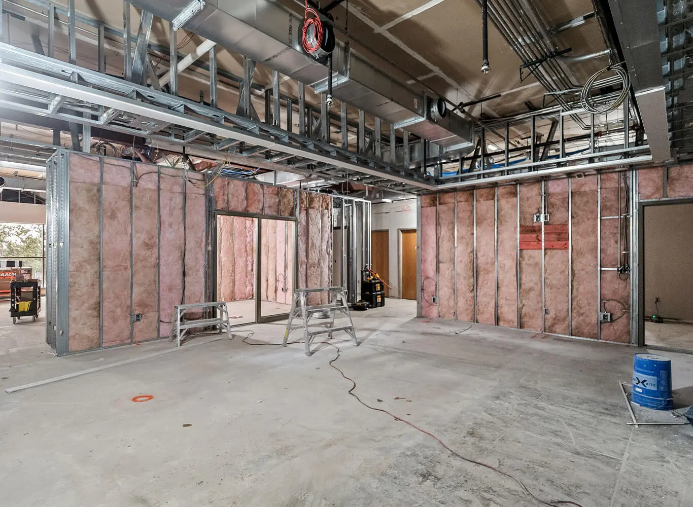 Interior of a large commercial building under construction, featuring exposed metal stud framing, pink batt insulation, overhead ductwork and conduit, and a concrete floor with a stepladder in the center.