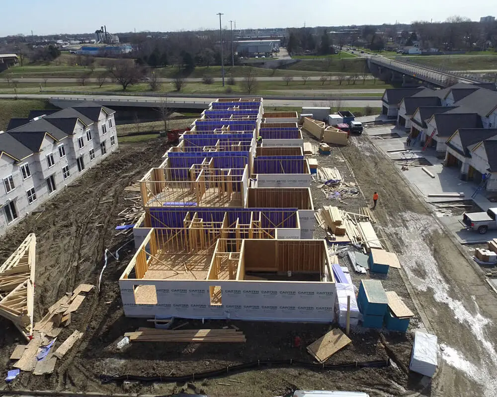 Aerial drone view of a multi-unit townhome or residential development under construction, showing wood framing and blue foam insulation across multiple units in a row, flanked by completed white-sided buildings and a highway overpass in the distance.