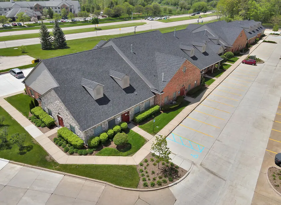 Aerial view of a completed commercial building complex with stone and brick façade, gray shingled roofs, manicured landscaping, and an adjacent parking lot with accessible parking spaces marked in blue.