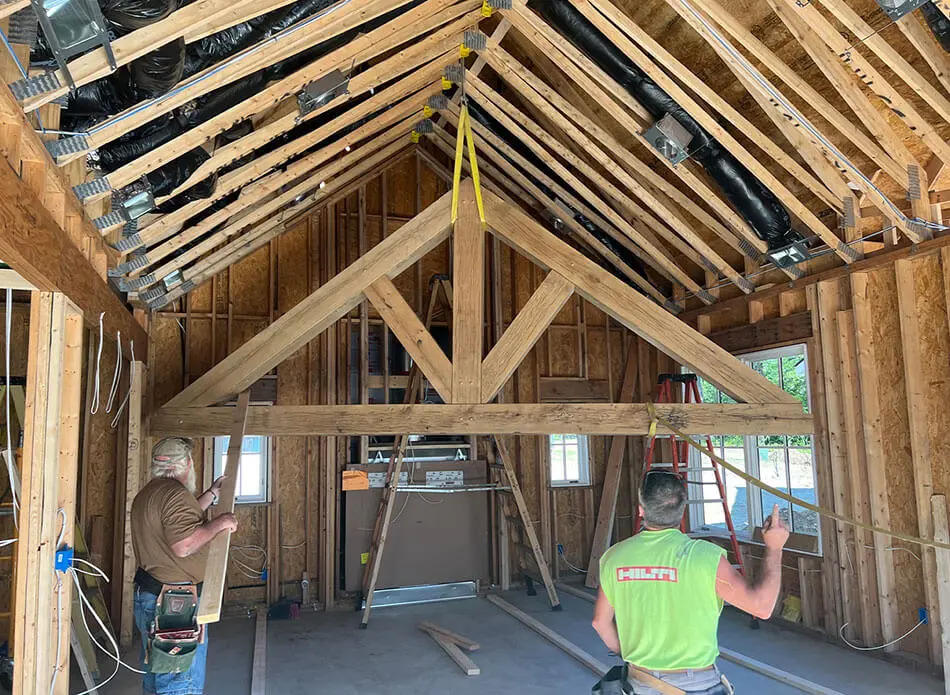 Interior view of a building under construction showing exposed wooden roof trusses and rafters with HVAC ductwork, while two workers assess a large timber beam being positioned in the open-concept framed space.