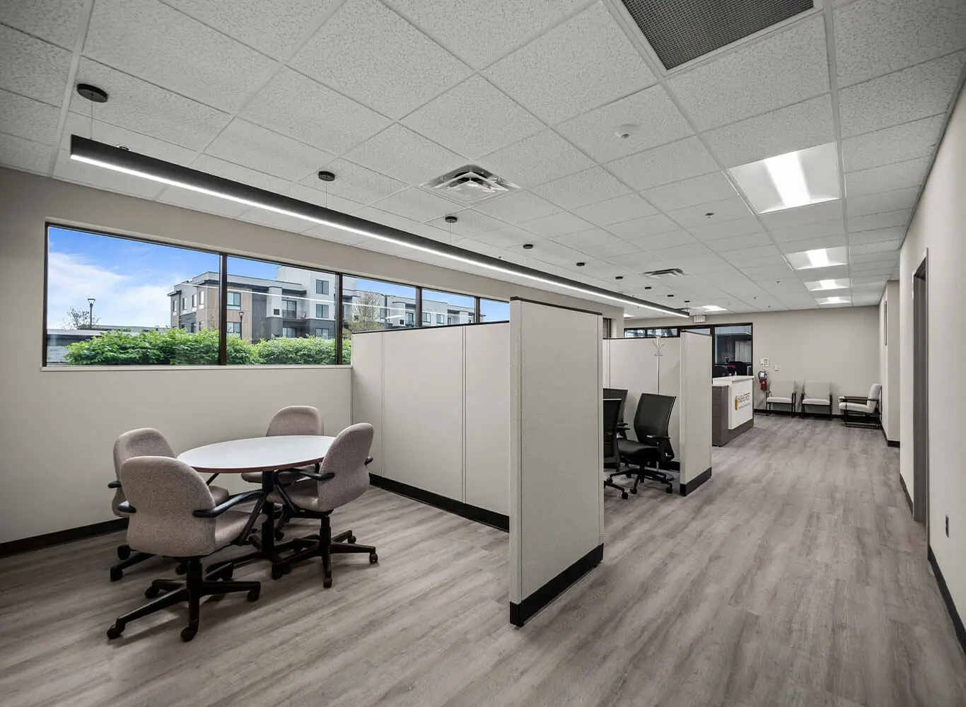 Interior of a newly built commercial office space featuring an open floor plan with cubicle dividers, a round meeting table, wood-look vinyl flooring, drop ceiling with recessed lighting, and a reception desk in the background.