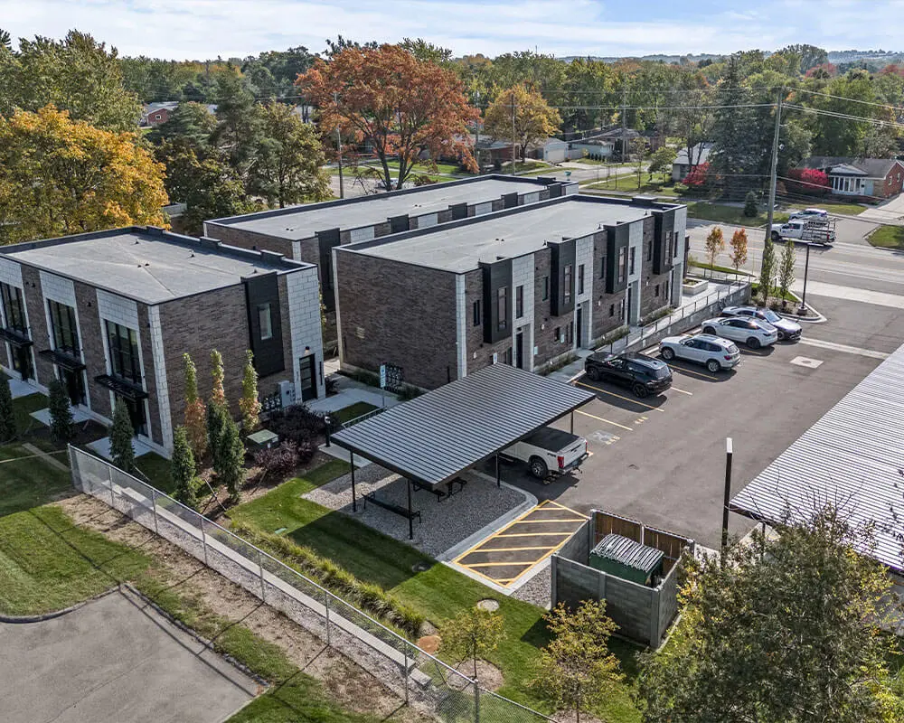 Aerial view of a modern multi-story residential townhome development with dark brick and panel exterior, flat roofline, parking lot, covered outdoor seating area, and fall foliage in the surrounding neighborhood.