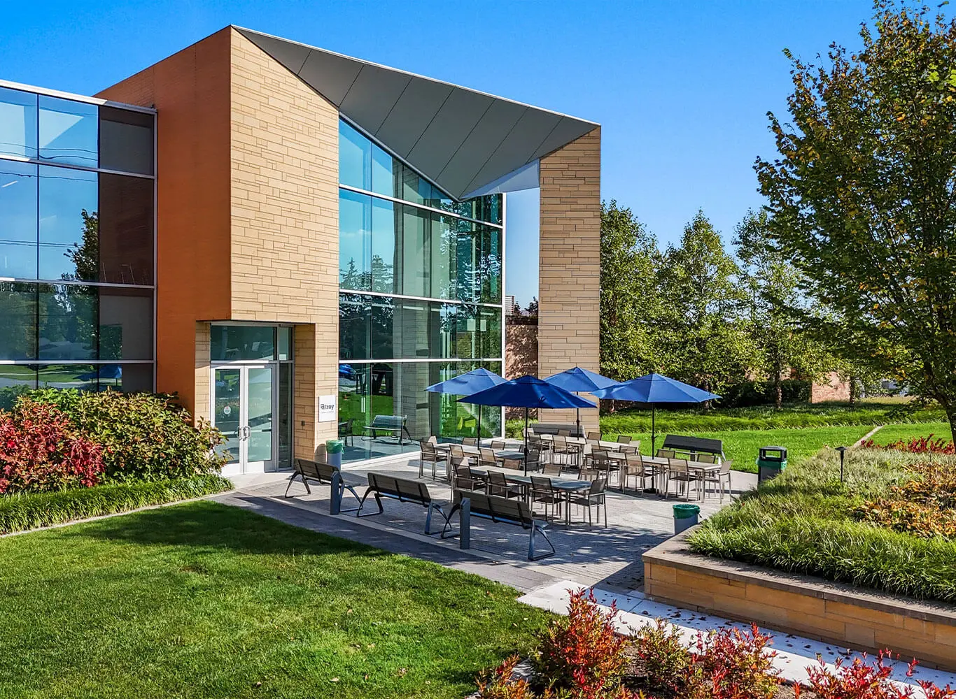 Ground-level exterior view of a modern commercial building entrance featuring floor-to-ceiling glass windows, tan brick columns, a dramatic angled canopy, outdoor dining tables and chairs with blue umbrellas, and lush green landscaping on a sunny day.
