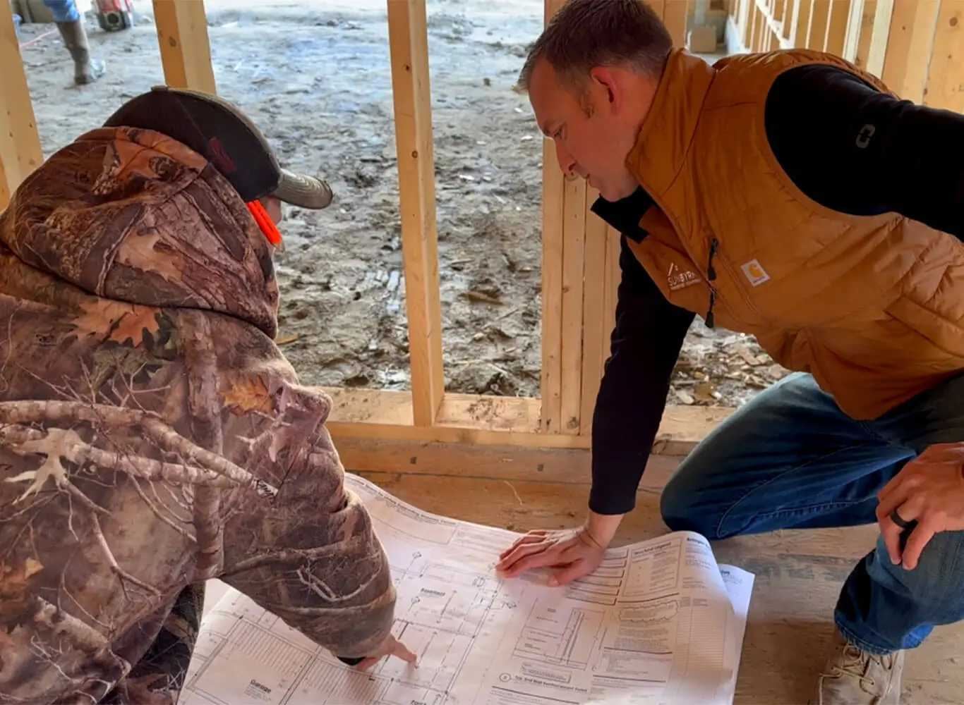 A SunByrnes team member in an orange vest crouches on a construction site floor to review blueprints with a colleague in camouflage gear, with wood framing and a muddy job site visible in the background.