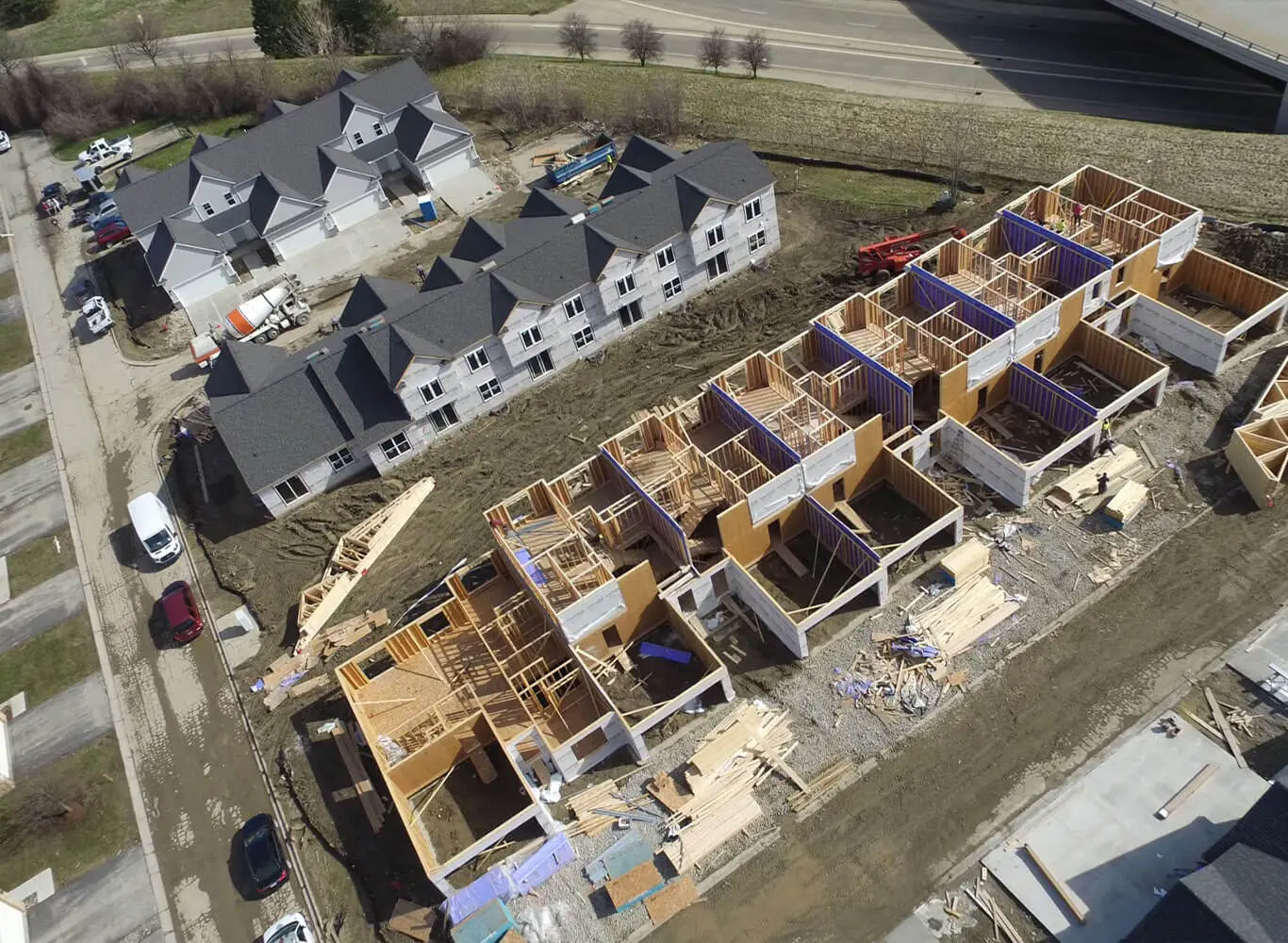 Aerial drone view of a residential construction site showing a row of townhomes in various stages of framing with exposed wood structure and blue insulation panels, alongside a completed multi-unit building with dark roofing, surrounded by construction equipment and materials