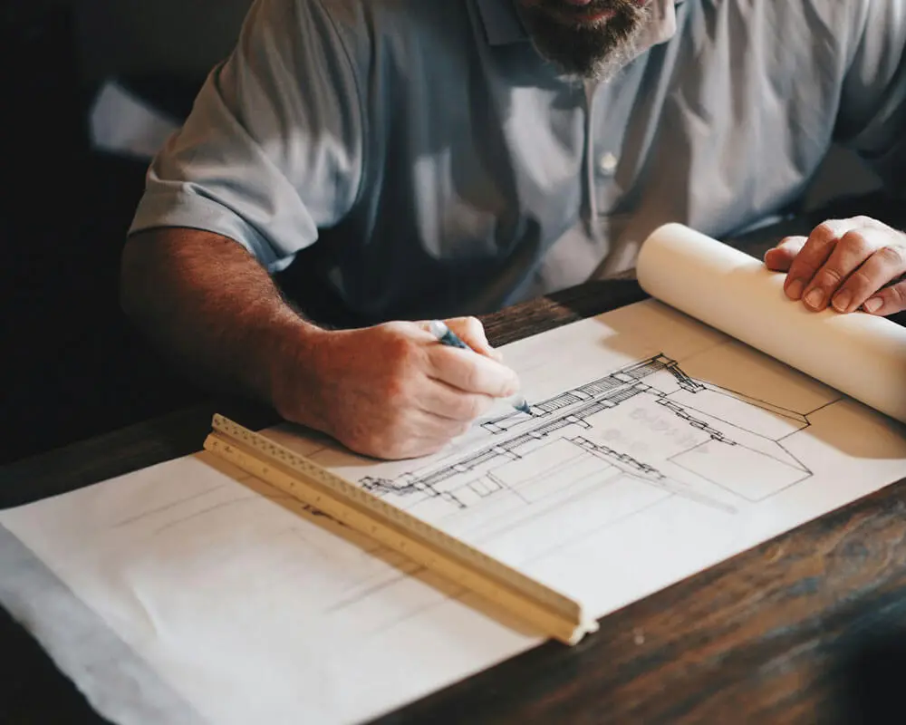 Close-up of a person's hands drawing architectural blueprints on drafting paper with a pen and wooden ruler, with a rolled blueprint in the background