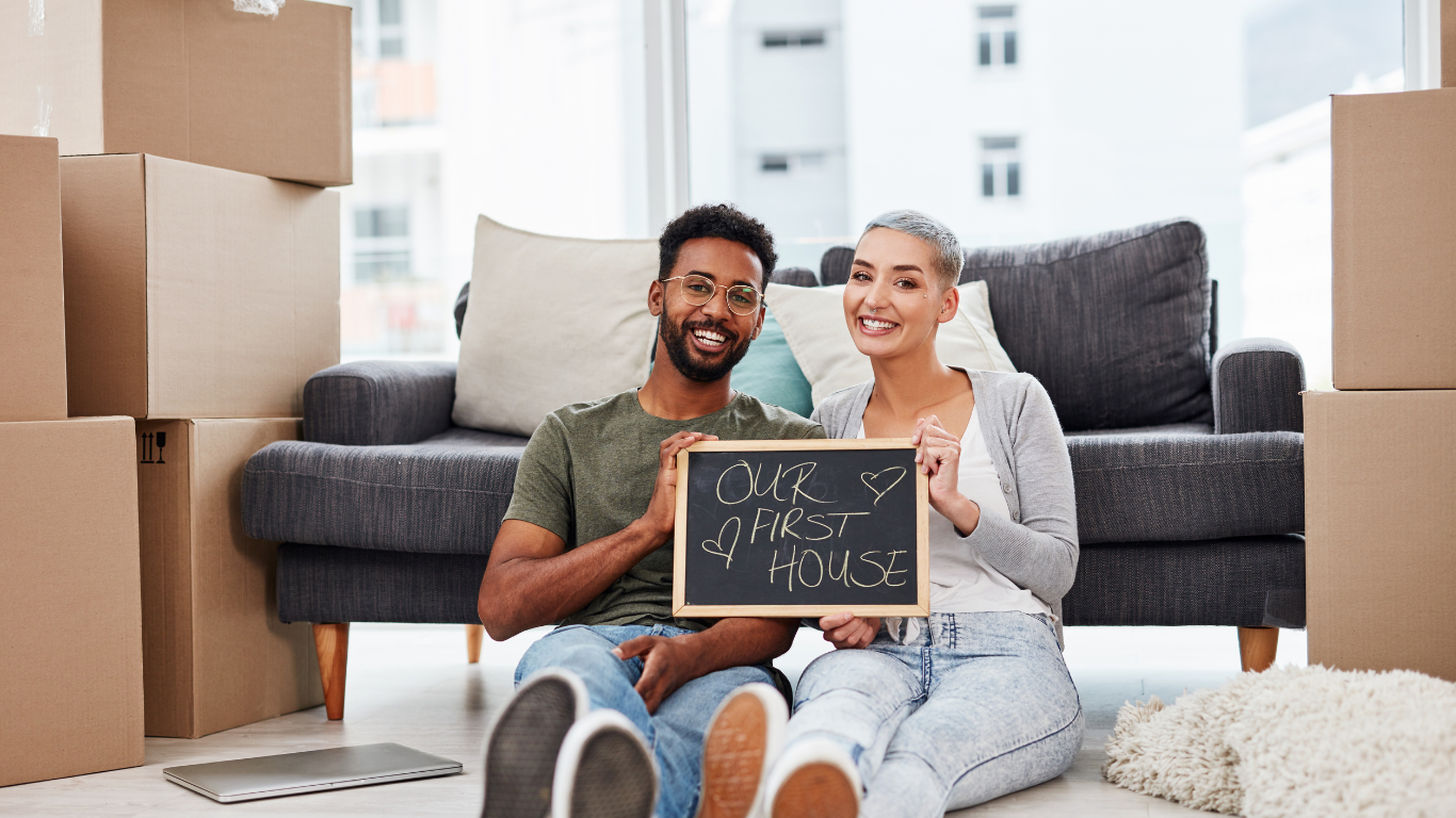 Happy first-time homebuyers sitting among moving boxes and holding a chalkboard sign that says “Our First House,” representing new homeowners in Michigan.